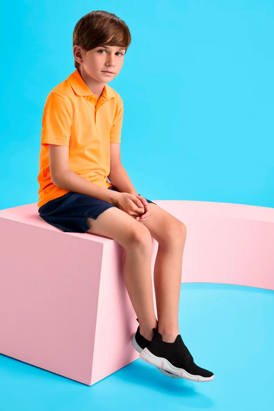 Poika istumassa yllään oranssi lasten pikeepaita ja tummansiniset shortsit.