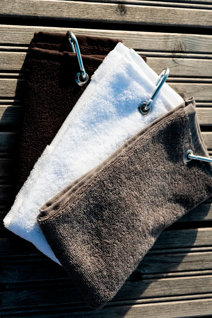 Kolme frotee-golfpyyhettä karbiinikoukulla ja metallirei'illä puisella pinnalla.